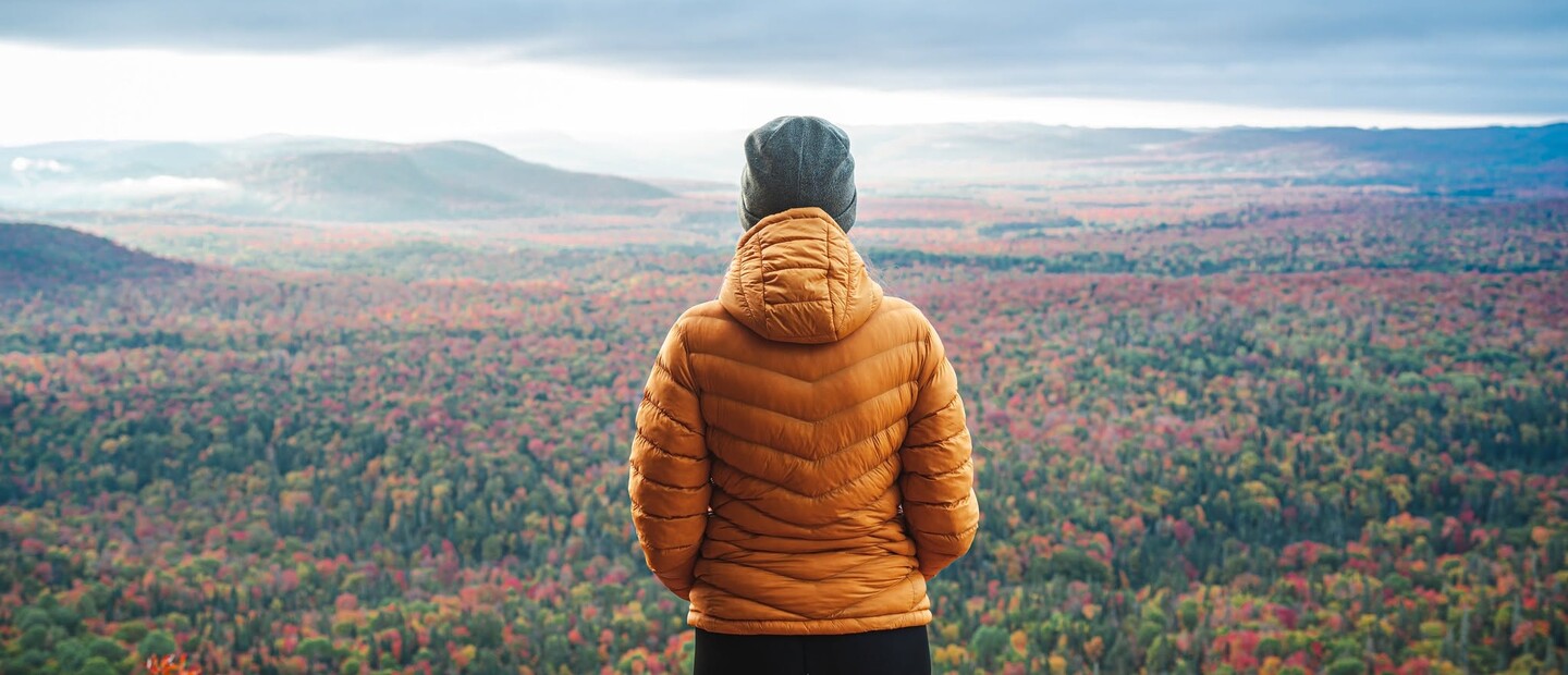 a person in a cozy jacket and toque stands on Robertson Cliffs on a cool autumn day, looking out over the dramatic view of the autumn forest and grey mist below.