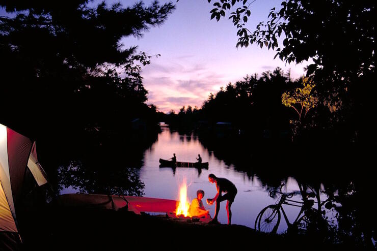 Lakeside campsite at dusk with tent, canoe, campfire and bicycles. 