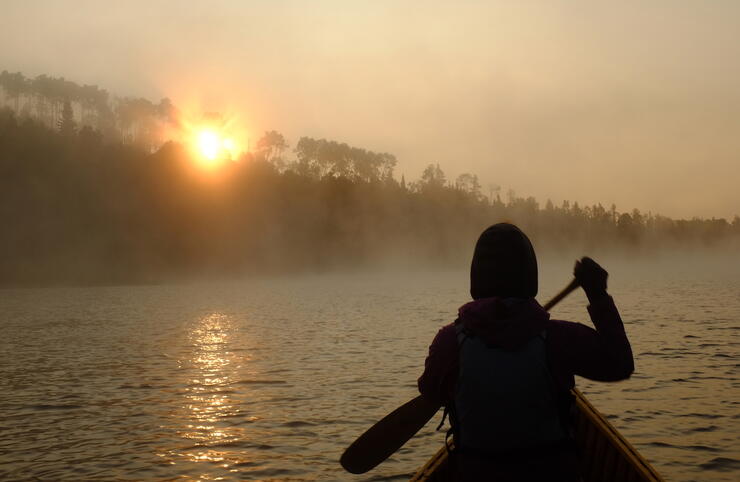 Canoeist paddling into sunrise on misty lake. 