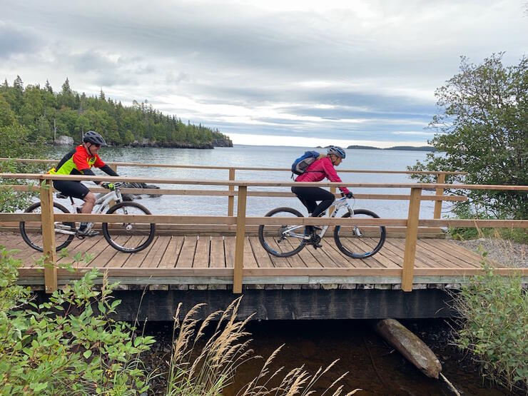 Two mountain bikers riding across a wooden bridge beside water.