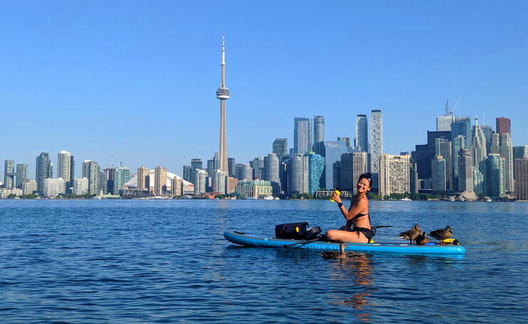 Young woman sitting on SUP board with ducks, Toronto skyline in background