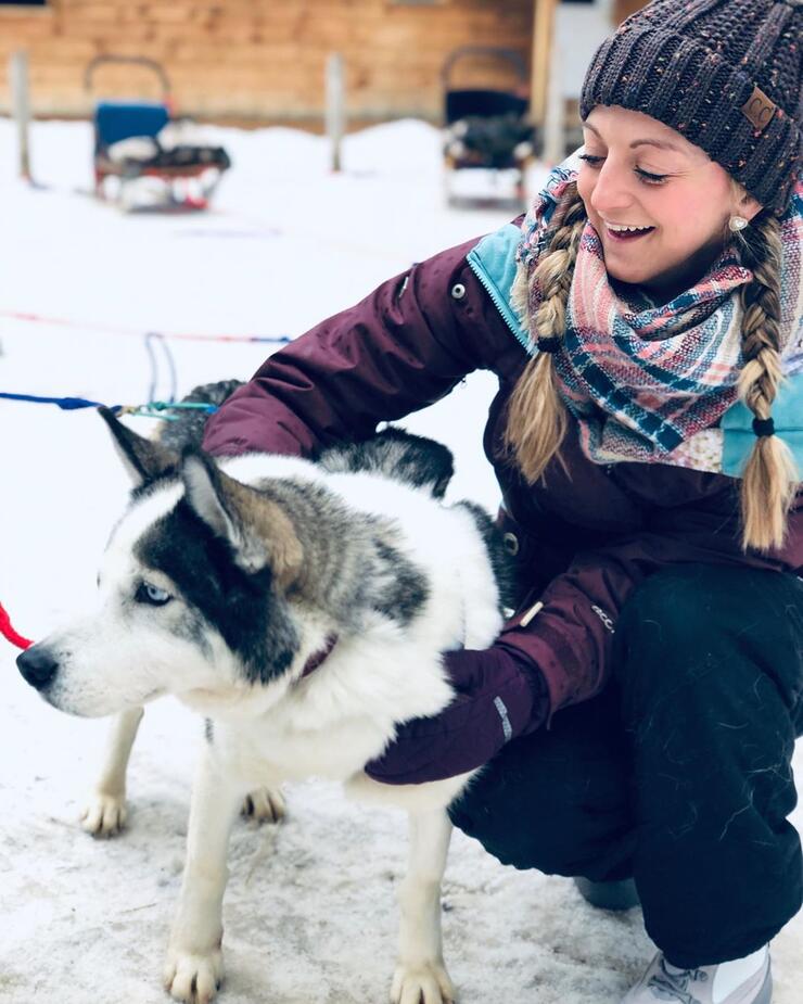 Young woman patting a blue eyed husky with dog sled in background. 