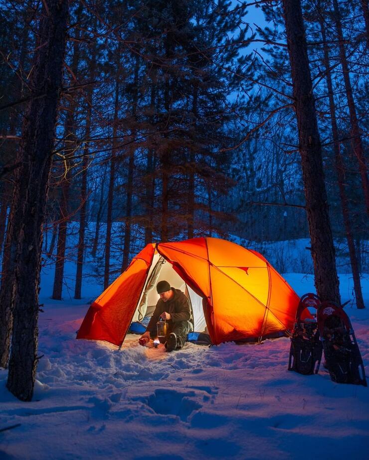 Man sitting in the door of an orange tent on a snowy campsite. 
