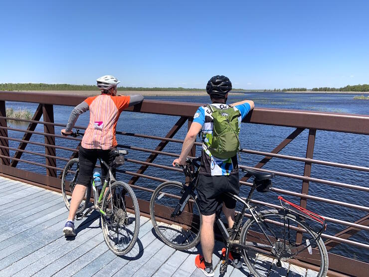 Two cyclists stopped on a wooden bridge looking out over water. 