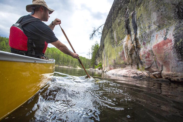 Man in a canoe paddling beside a rock face with pictographs 