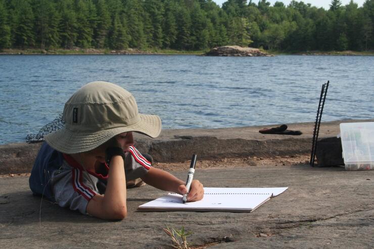 Child writing in a journal while laying on a rock beside a river