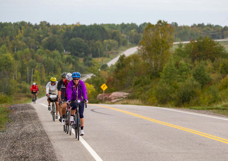 Group of cyclists riding in single file on shoulder of paved road. 