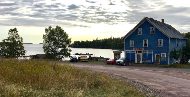 Looking a front of a A-frame structure with a gravel road in front and a lake beside it. 