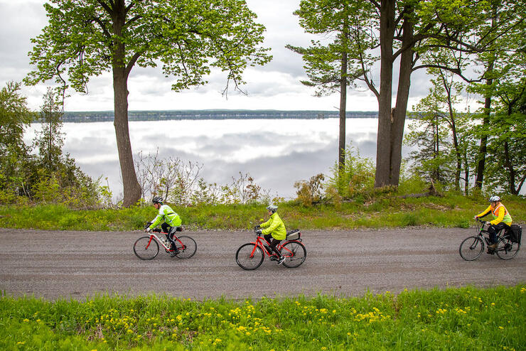 Three cyclists riding on a flat gravel road beside a lake and trees