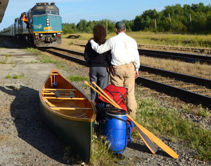 Man and woman standing beside canoe and gear while train approaches.