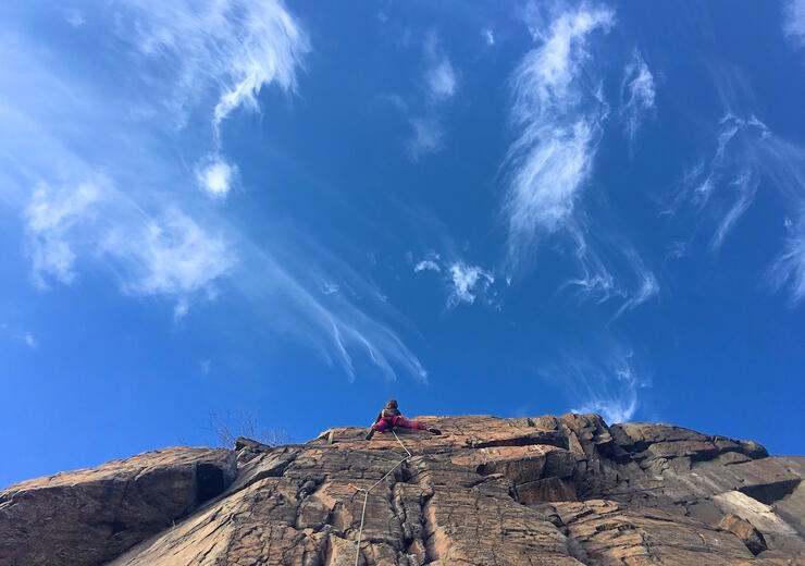 Person climbing half way up tall rock face with blue sky and wispy clouds.