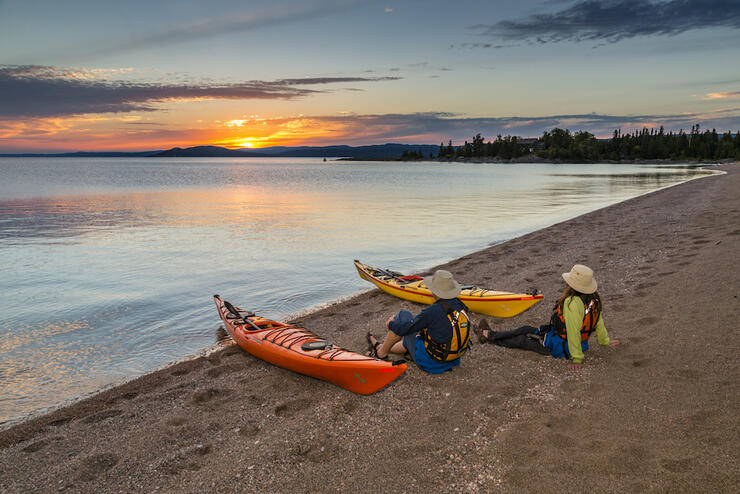 A couple sitting on sandy beach beside kayaks with sun setting over water
