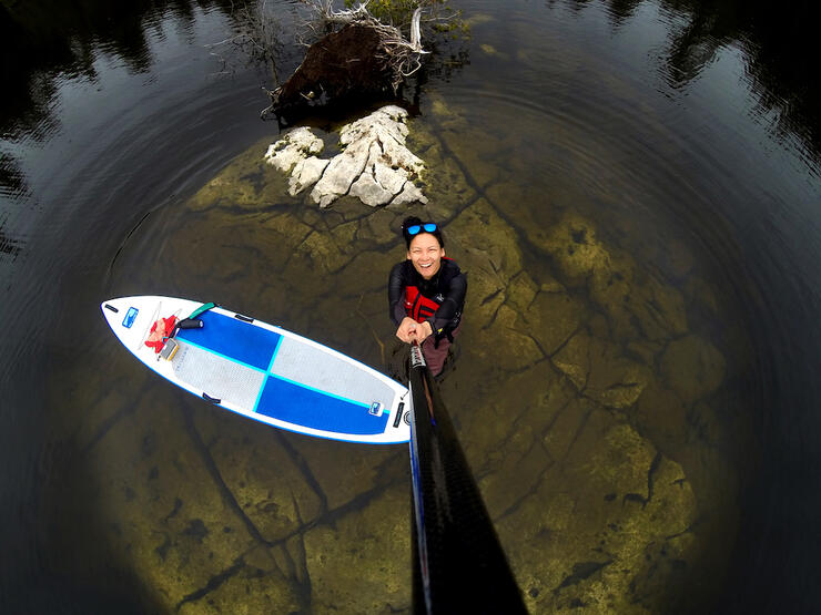Young woman taking a selfie with paddle looking down at SUP, rocks and water. 