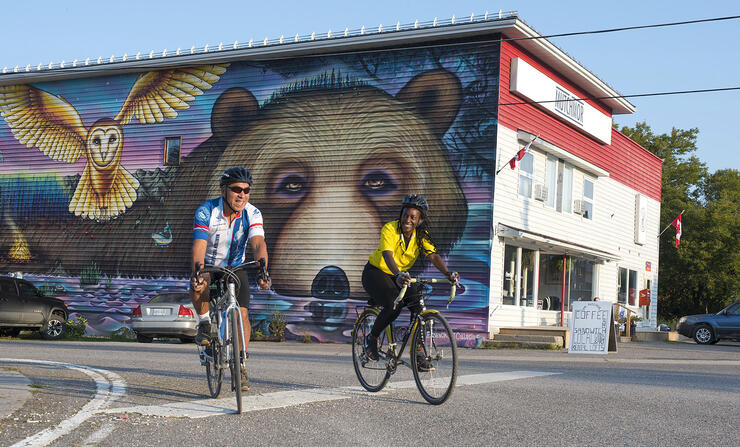 Two cyclists riding past a building painted with a mural of bear and owl.