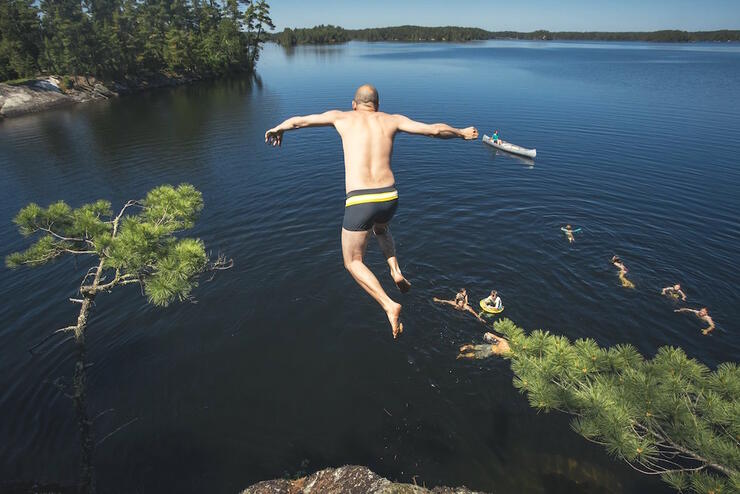 Man jumping off rock cliff into lake with several other swimmers