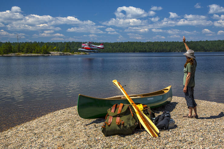 Woman on beach with a canoe and gear looking at a floatplane taking off in the distance
