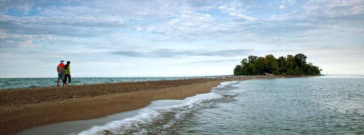 Couple walking on a sand spit towards tree covered island.