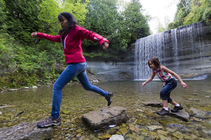 A girl and woman jumping across rocks in river with falls in background. 