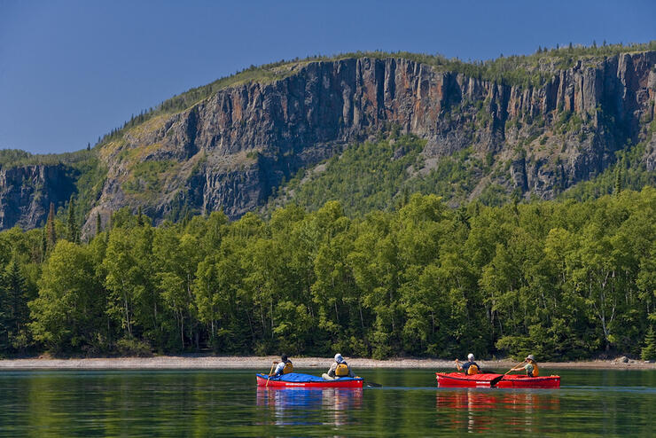 Two canoes paddling on calm water infront of large rock cliff.
