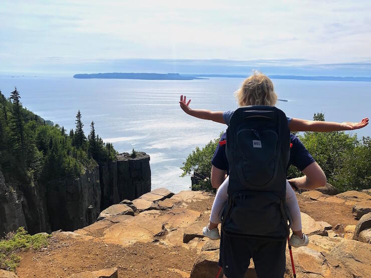 Person with young child in back carrier look at view of Lake Superior from high cliff
