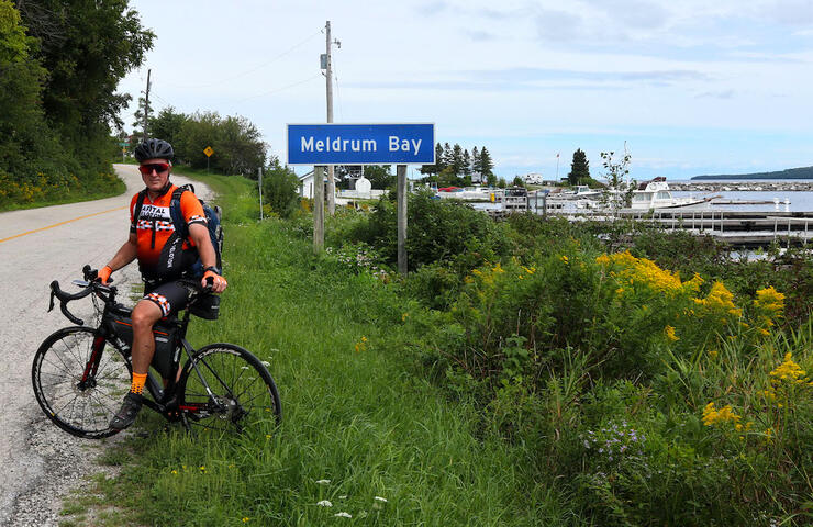 Man on a bicycle in front of Meldrum Bay road sign and marina 