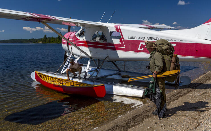 Man with pack and paddles standing near floatplane with canoe. 