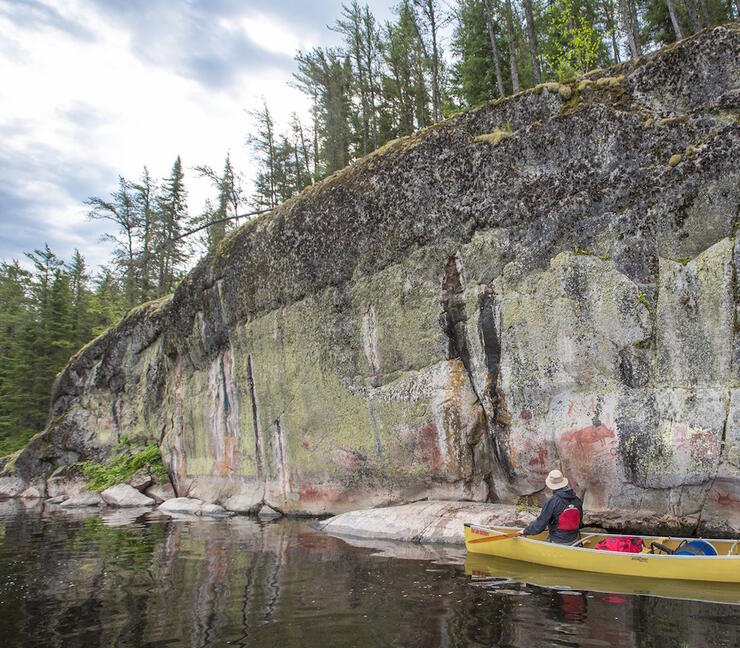 Man in a canoe paddling along a rock face covered in ancient pictographs.