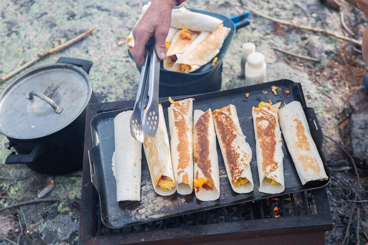 Rolled wraps cooking on a griddle over a campfire. 