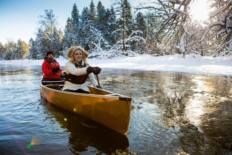 Woman and man in winter clothing paddling a yellow canoe along snow-covered shoreline