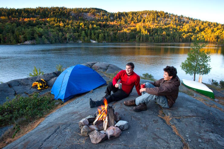 Two men sitting by a campfire overlooking lake - tent and canoe in background.