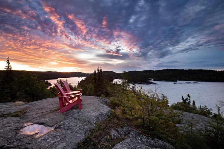 Two red Muskoka chairs perched on top of a top overlooking sunset on Lake Superior.