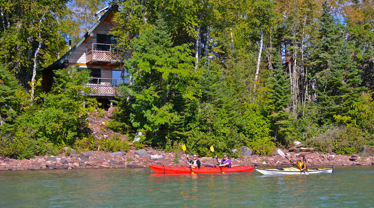 Kayakers paddling by a log cabin. 
