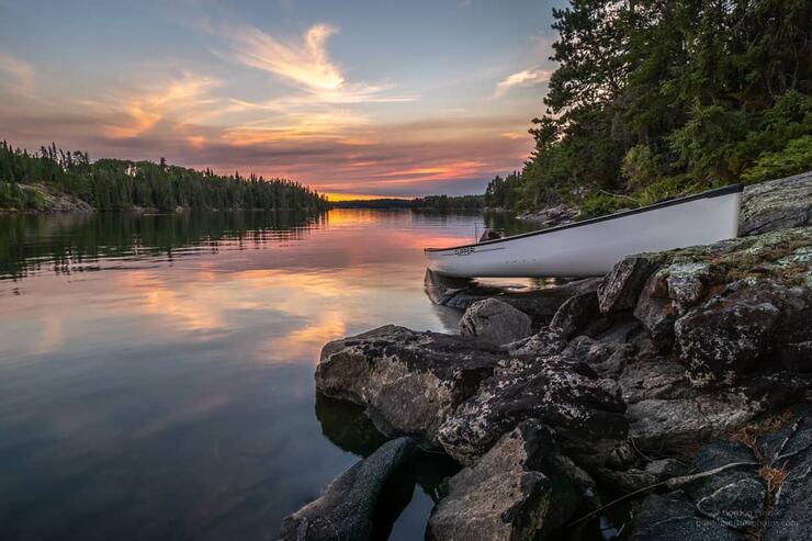 Empty white canoe pulled up on shore beside a calm river at sunset. 