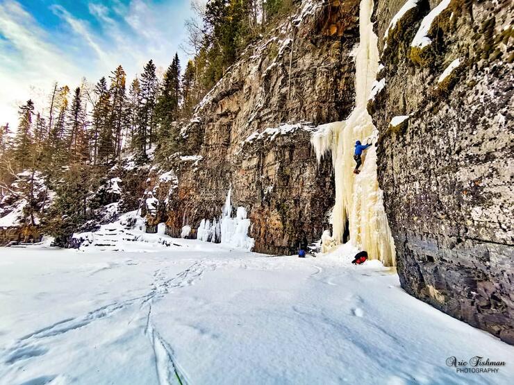 A climber ascending an ice covered rock face  beside a snow covered  lake. 