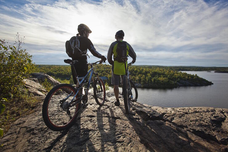 Two cyclists standing with bikes on a rocky lookout overlooking a lake. 
