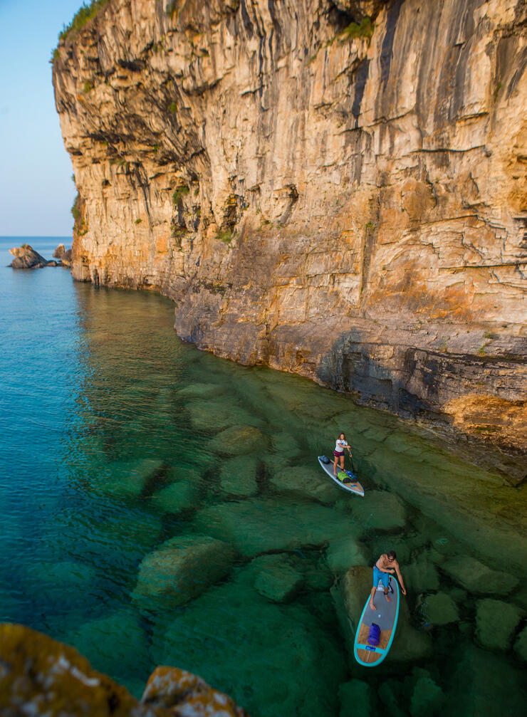 Two people stand-up paddle boarding along side tall cliffs 