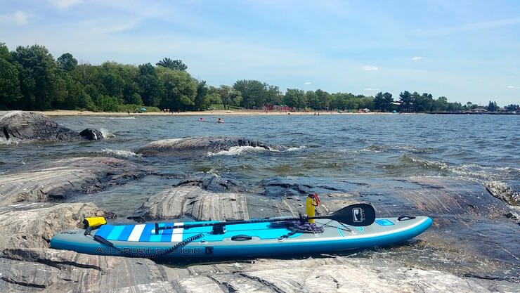 Blue paddleboard on flat rocks beside lake with beach in background