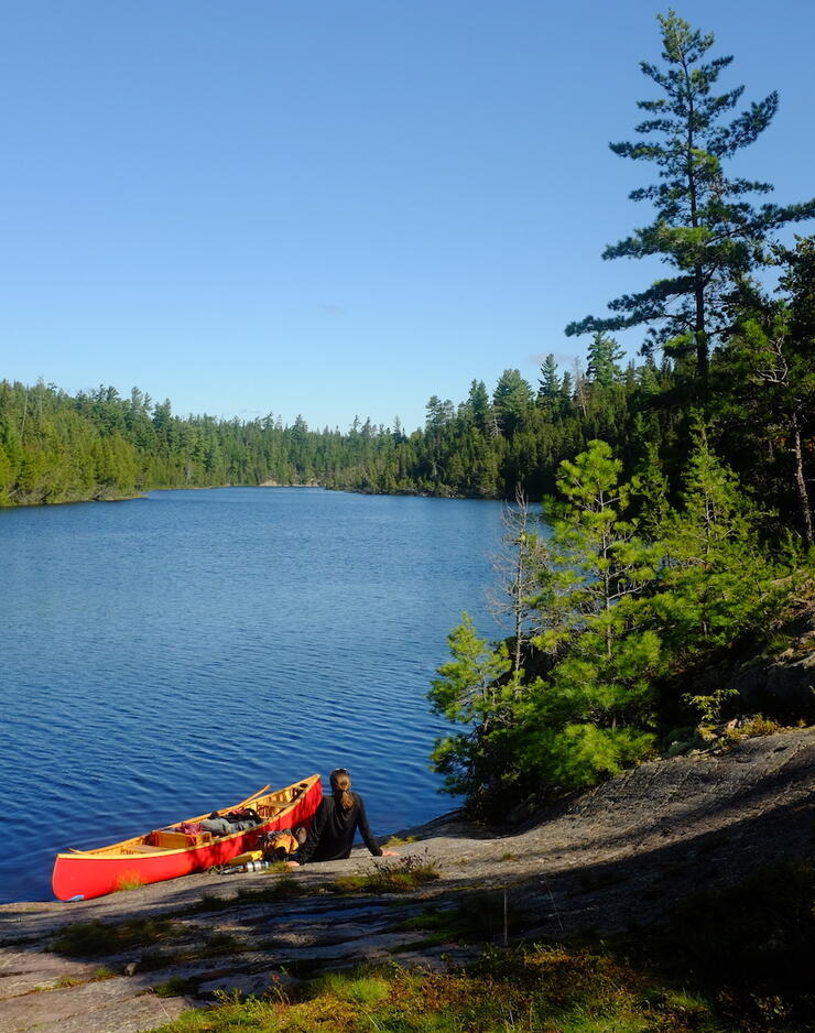 Woman sitting on smooth rock shore with red canoe, looking down a lake