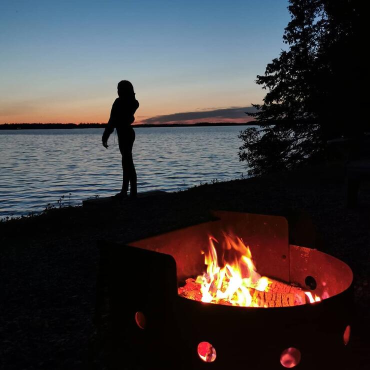 Woman standing on the shore of a lake at sunset with a campfire in foreground.