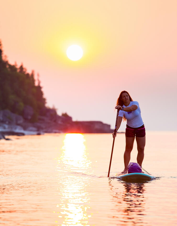 Strong woman stand-up paddle boarding in front of warm sunset 