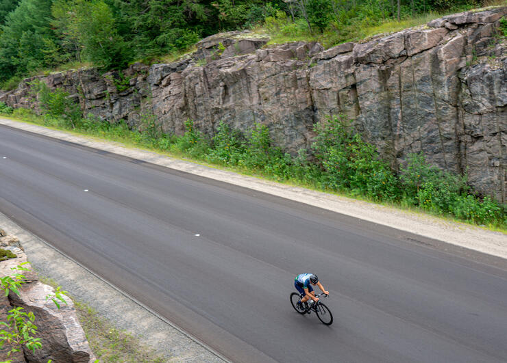 Man on a bike riding on a two lane highway beside large granite rockface