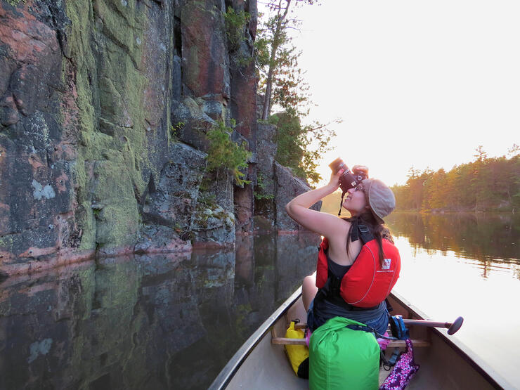 Woman photographing rocky cliffs while in a canoe on a river.