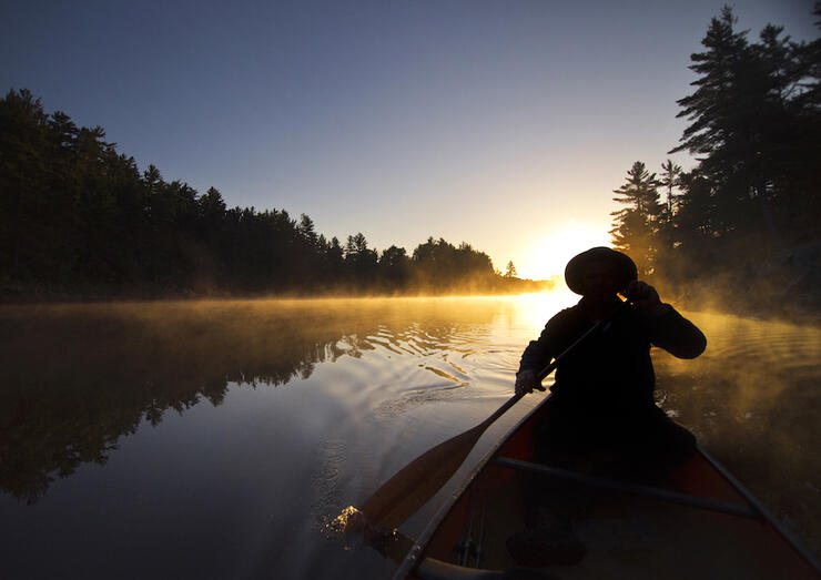Canoeist in bow of canoe paddling into misty sunrise 