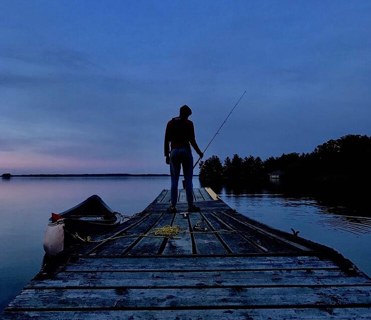 Man standing at the end of a dock fishing at twilight.