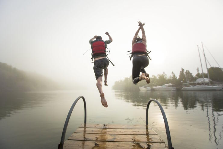 Two people in life jackets jumping off end of dock into water on a misty morning.