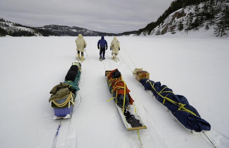 Three people, each pulling a toboggan packed with winter camping gear, on a frozen lake