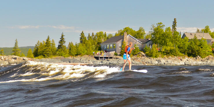 Woman on SUP in front of small lodge on rocky point. 