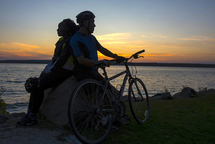 Man and woman sitting back to back holding a bicycle, at sunset.