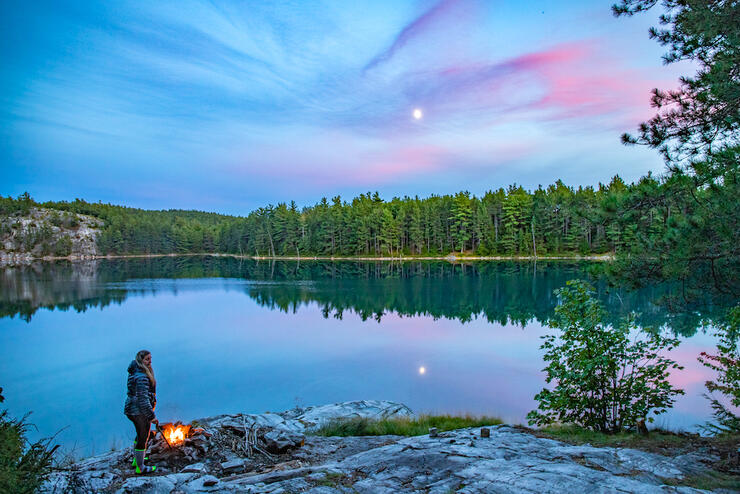 Woman standing beside a campfire looking at beautiful lake with pink swirls in sky.