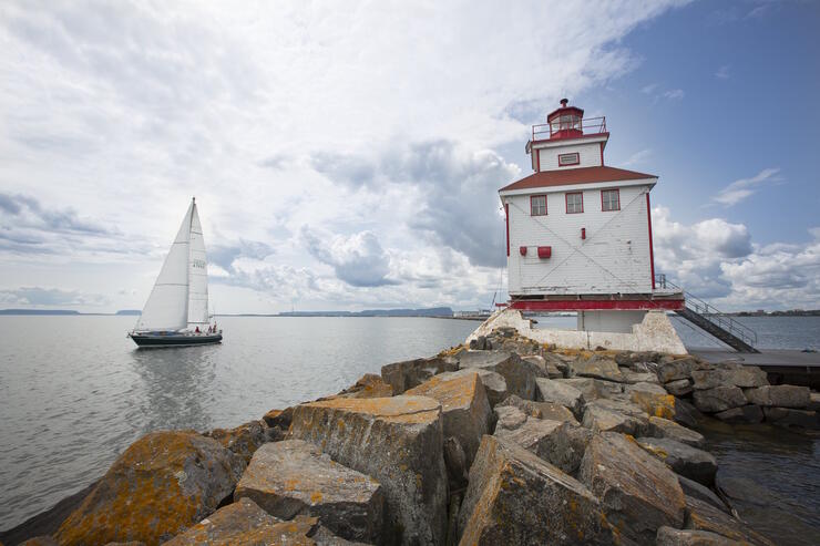 Lighthouse on rocks with a sailboat sailing by. 